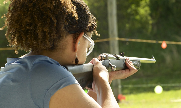 photo depicts a woman from the back pointing a rifle towards a target outdoors