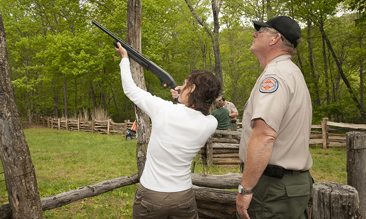 photo depicts a woman in a white shirt outdoors pointing a shotgun upward as a hunter education instructor in uniform stands nearby looking up